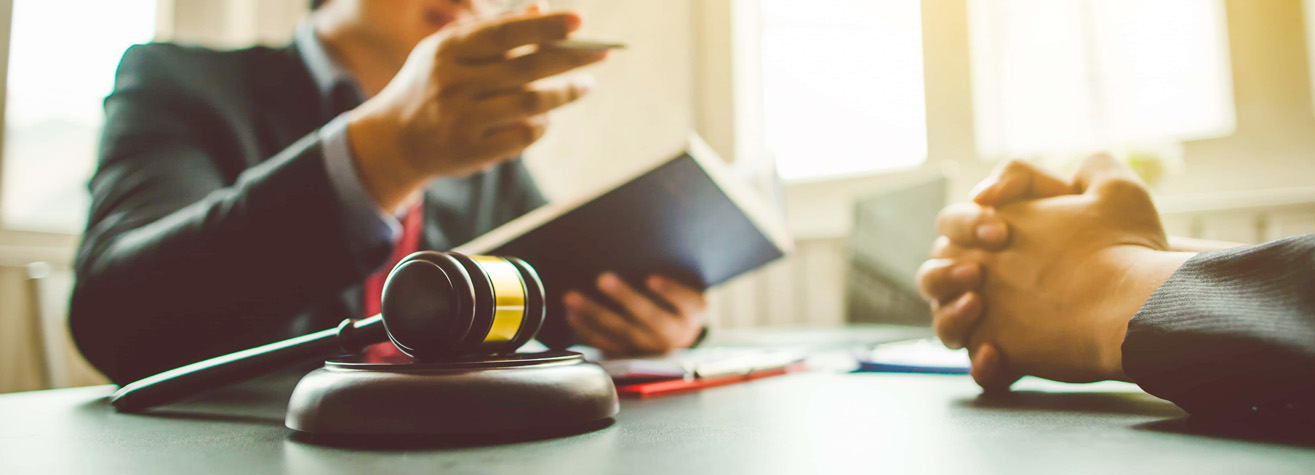 A close-up of a desk with a gavel in the foreground. In the background, two people are engaged in conversation. One is holding a book, and both are wearing suits, suggesting a legal or professional setting.