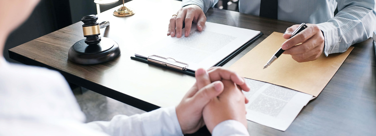 Two people are sitting across from each other at a wooden desk. One person is holding a pen and gesturing towards a document, while the other person has their hands clasped. A gavel and a lamp are on the desk.