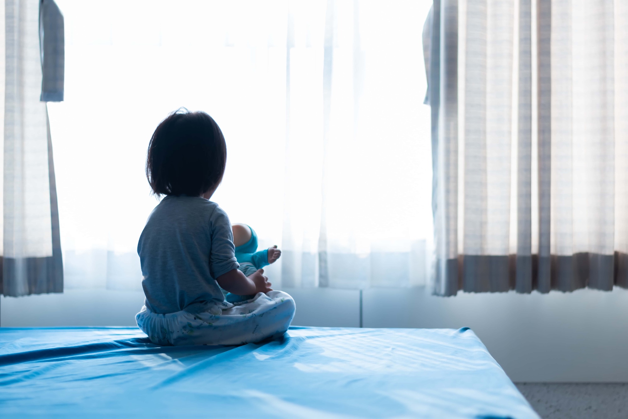 A child with short dark hair sits on a blue mat, facing away, holding a doll. The child is looking towards a brightly lit window with sheer curtains. The scene is serene and softly lit.