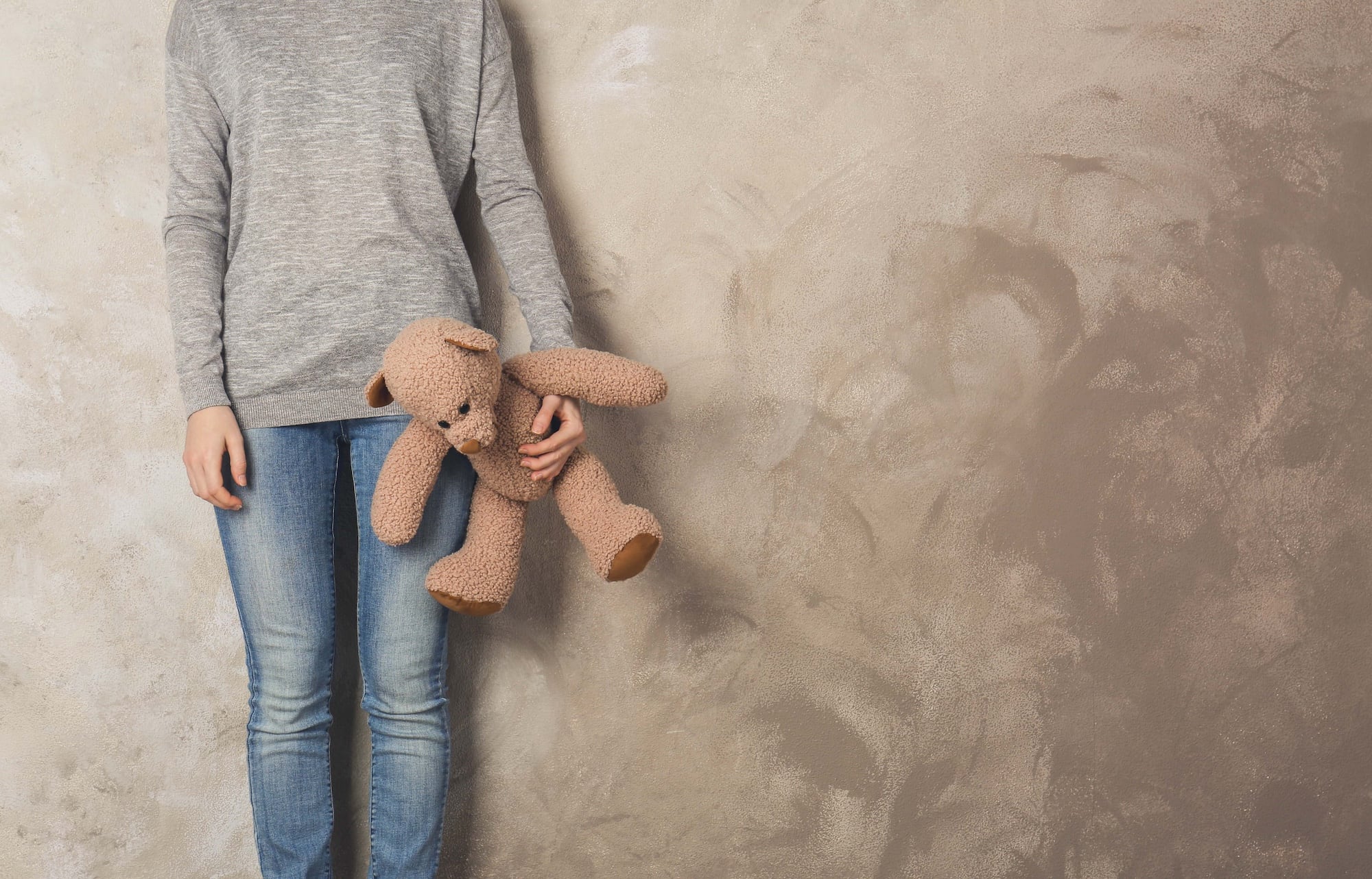 Person in a gray sweater and jeans holding a brown teddy bear by the arm, standing against a textured beige wall.