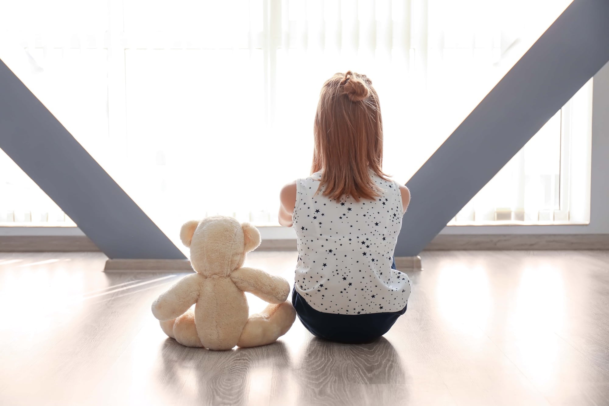 A young girl with shoulder-length hair sits on a wooden floor, facing a bright window. She wears a sleeveless top with a star pattern. Beside her is a teddy bear, also facing the window. Sunlight streams in, creating a serene atmosphere.