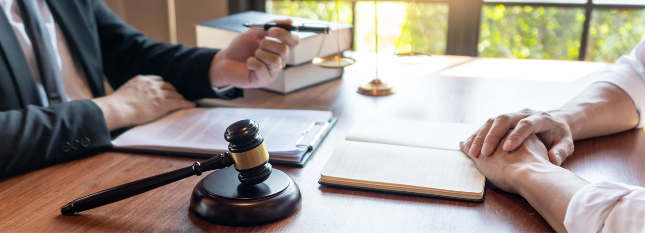 Two people sit at a wooden desk, one in a suit gesturing with a pen over a clipboard. A gavel rests nearby, and books are stacked on the table. Sunlight filters through a window, creating a calm atmosphere.