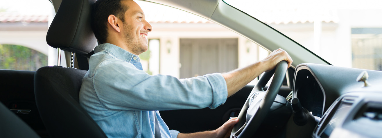A man is smiling while driving a car. He is wearing a light blue denim shirt and has short hair. The interior of the car is visible, showing the steering wheel and dashboard. A building is blurred in the background through the window.