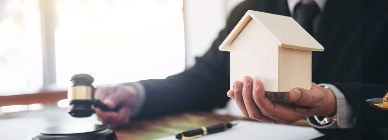 A person in a suit holds a small wooden house model in one hand and a gavel in the other, suggesting a legal or real estate context. A pen lies on the table in front of them.