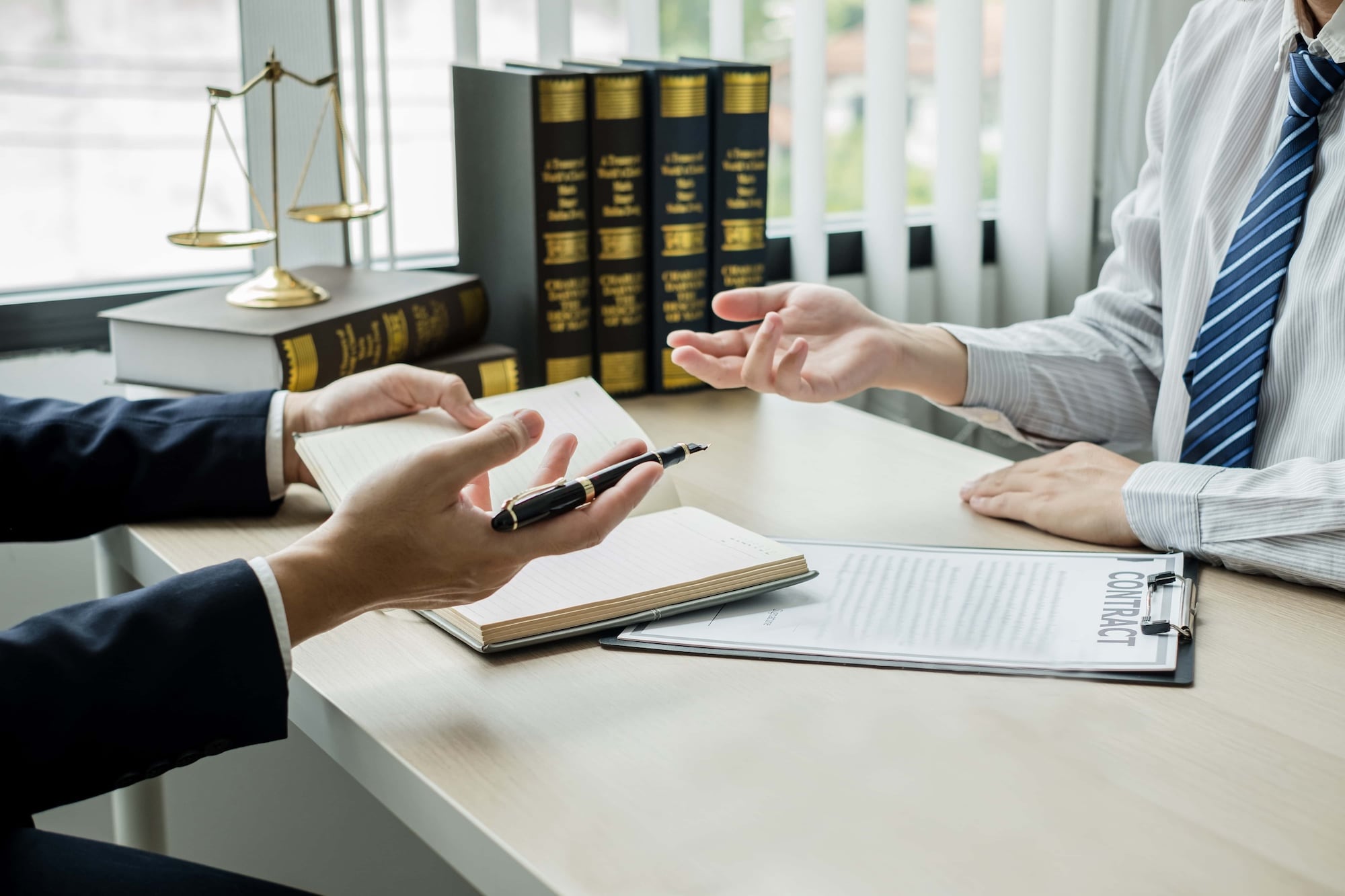 Two people in business attire are sitting at a desk, discussing over an open notebook and a contract. One holds a pen, gesturing towards the notebook, while the other listens attentively. Law books and a scale are visible in the background.