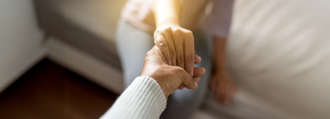 Close-up of two people holding hands. One person is reaching out with a gentle grip, and the other is seated on a couch, partially visible. The scene conveys a sense of comfort and support. The light is soft and warm.