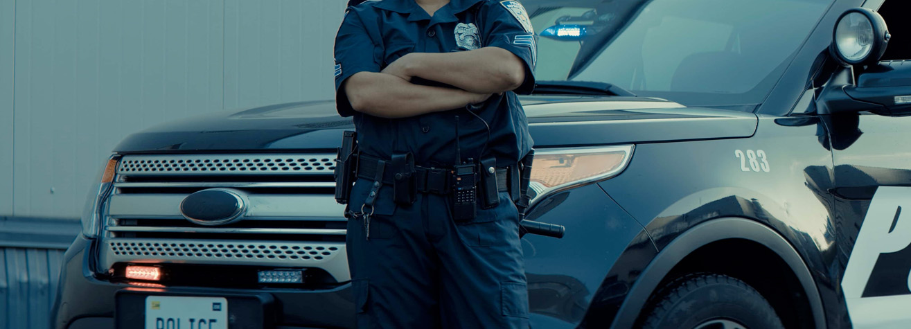 A police officer stands confidently with arms crossed in front of a dark-colored police SUV. The vehicle has its lights on, and the officer wears a uniform with various tools on the belt.