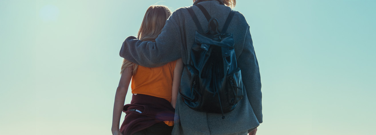 Two people stand close together, viewed from behind against a clear blue sky. One person has their arm around the other's shoulders. They are wearing casual clothing and a backpack.