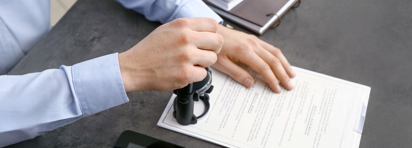A person in a blue shirt seals a document with a stamp on a clipboard placed on a dark table. Nearby are other papers and documents.