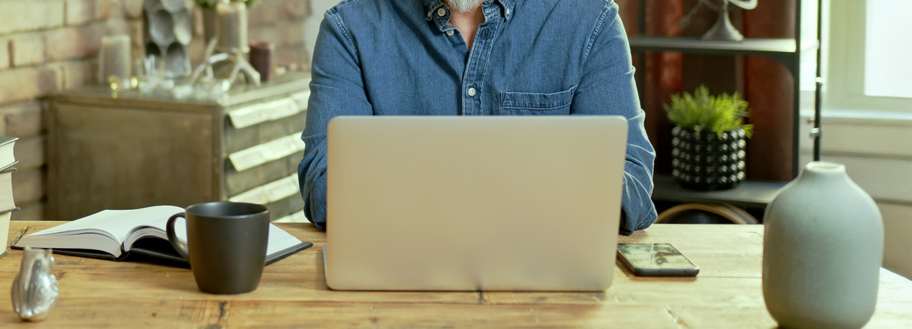 A person in a blue denim shirt sits at a wooden table, using a laptop. An open notebook, black coffee mug, smartphone, and a decorative vase are also on the table. In the background, shelves with plants and decor items are visible.