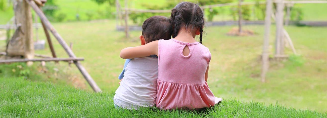 Two children sit on a grassy hill, seen from behind. The older child in a pink dress gently wraps an arm around the younger one in a white shirt. They gaze at a blurred vista of grassy fields and wooden structures, carefree under the watchful care akin to abogados de defensa de peligro infantil.
