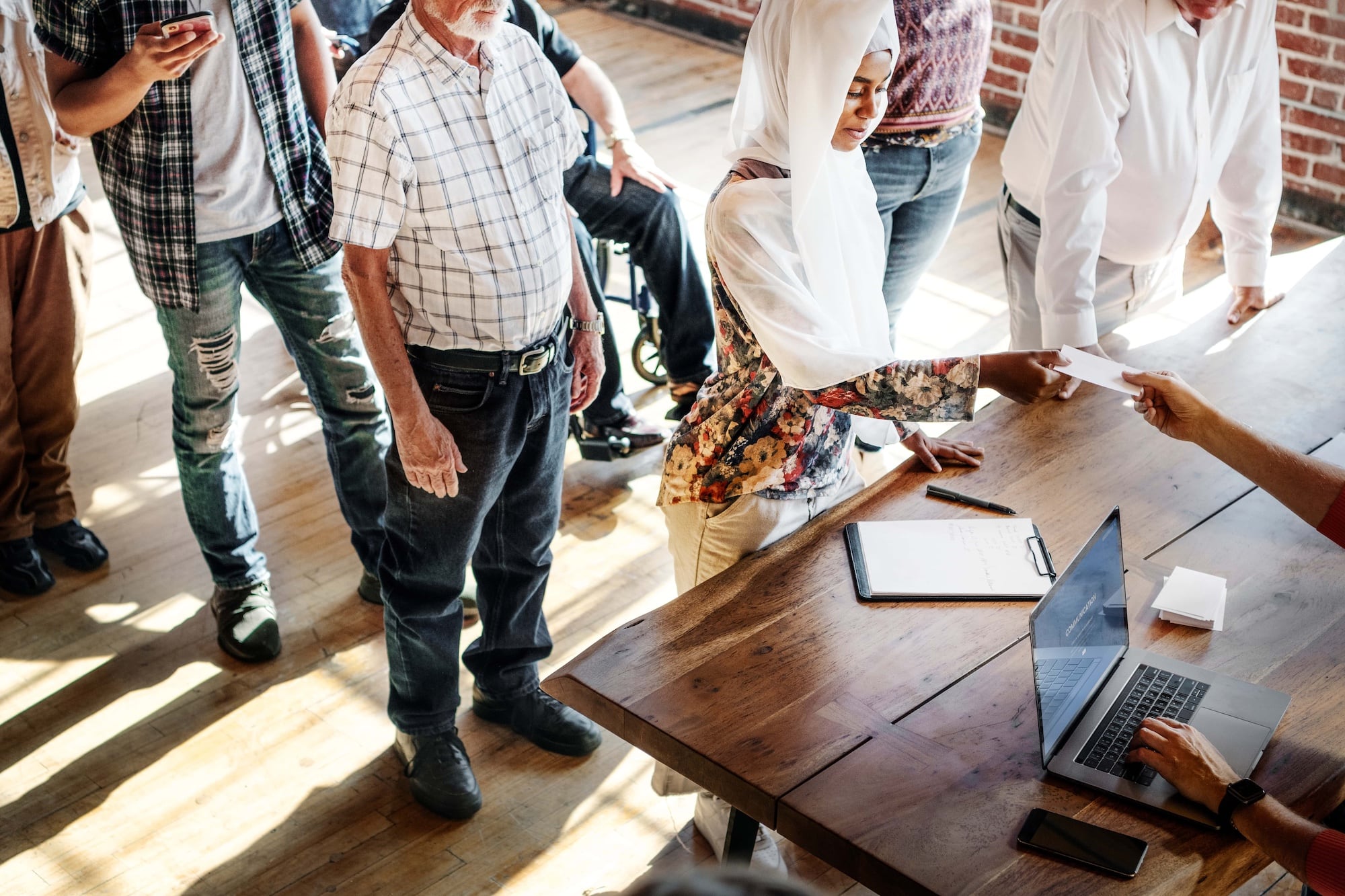 A diverse group of people stands in line at a wooden table where a person hands out papers. A laptop and clipboard are on the table. Sunlight filters through large windows, illuminating the scene.