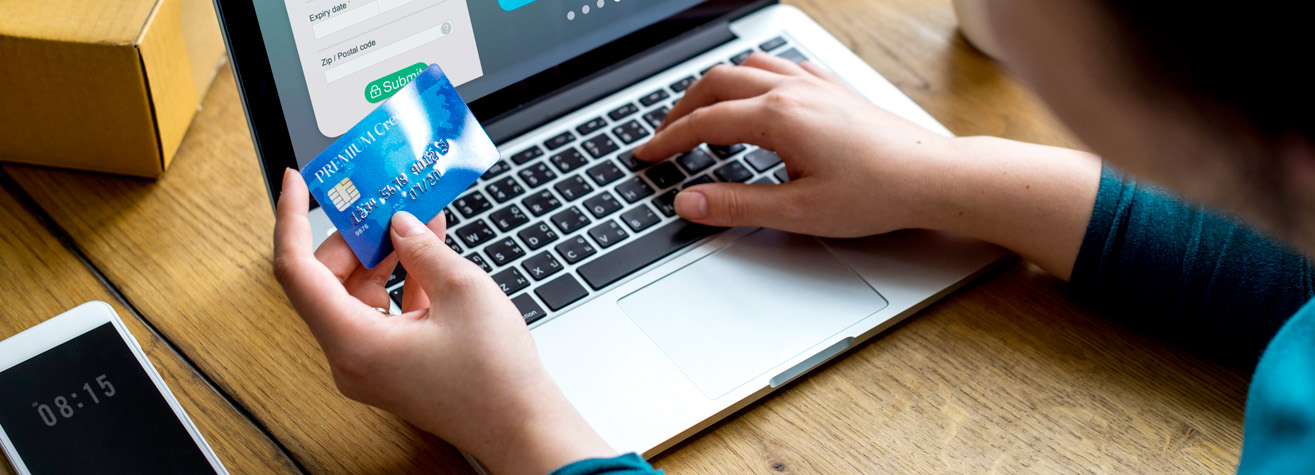 Person using a laptop for online shopping, holding a credit card in one hand. A brown box and smartphone with a black screen displaying 