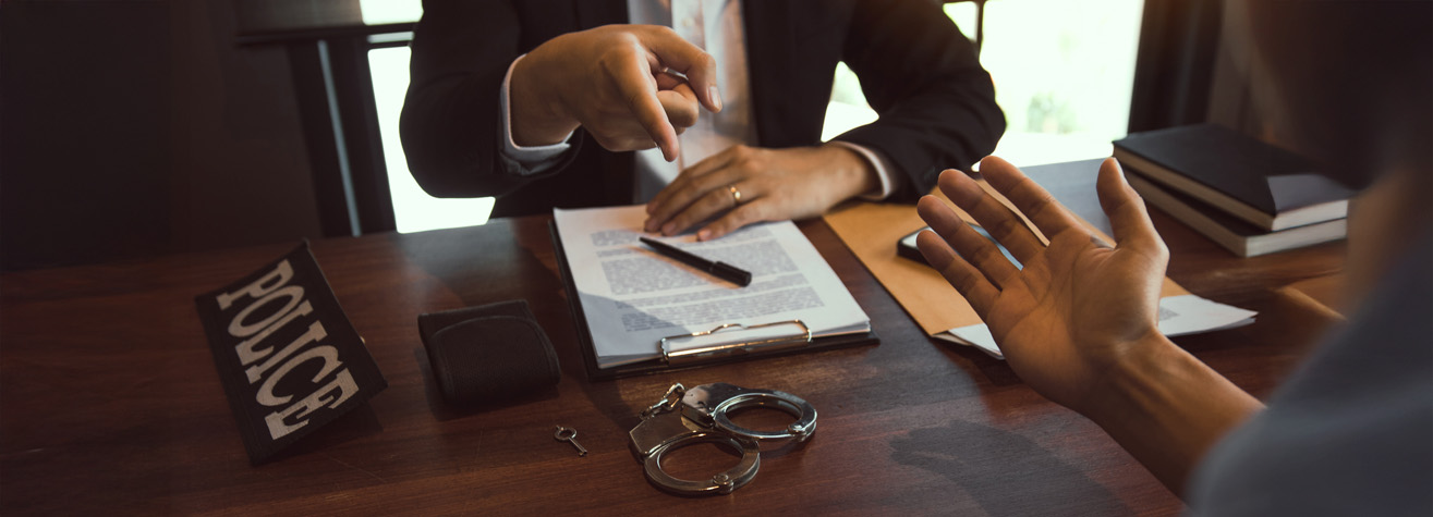 A person in a suit sits at a desk with documents, pointing at someone off-camera. A 