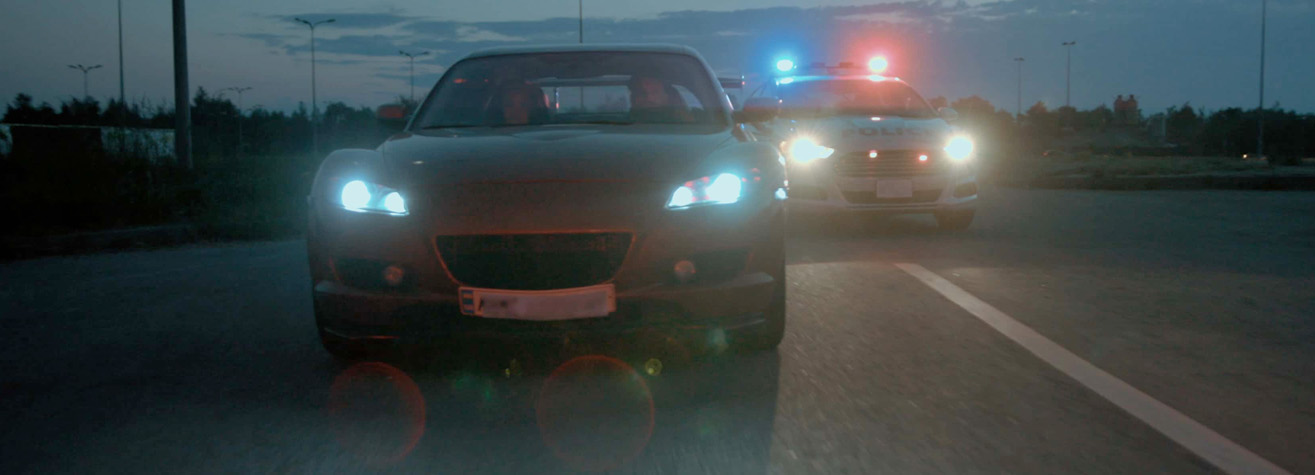 A car is being chased by a police car with flashing red and blue lights on a dimly lit road. The sky is dark, and the area appears to be deserted, adding to the tense atmosphere of the scene.
