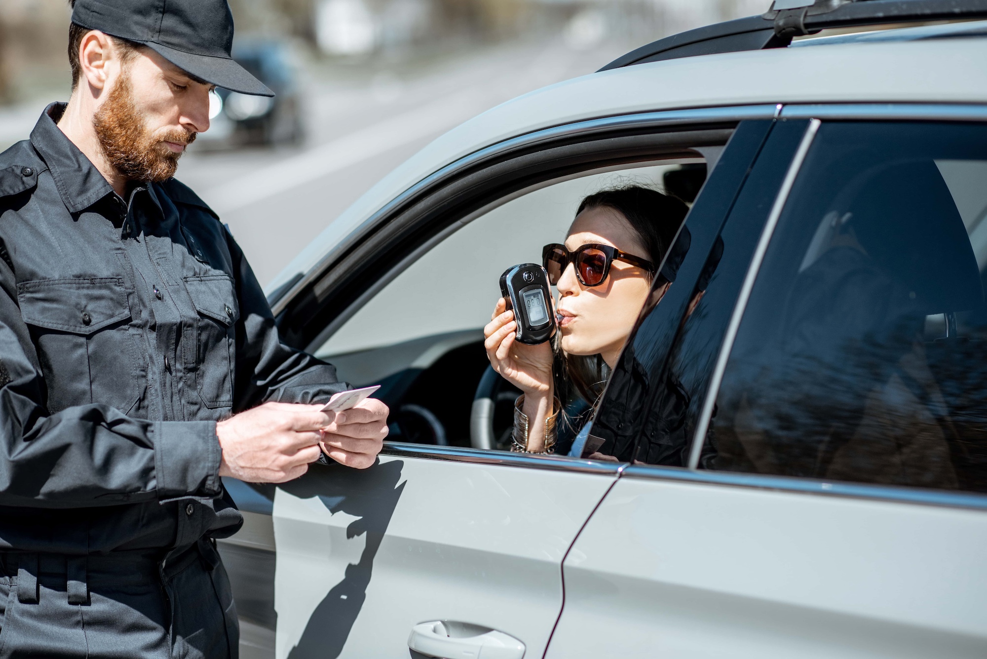 A woman in sunglasses sits in a car, using a breathalyzer. A uniformed officer stands beside the vehicle, holding a document and wearing a cap. The scene takes place on a road during the day.