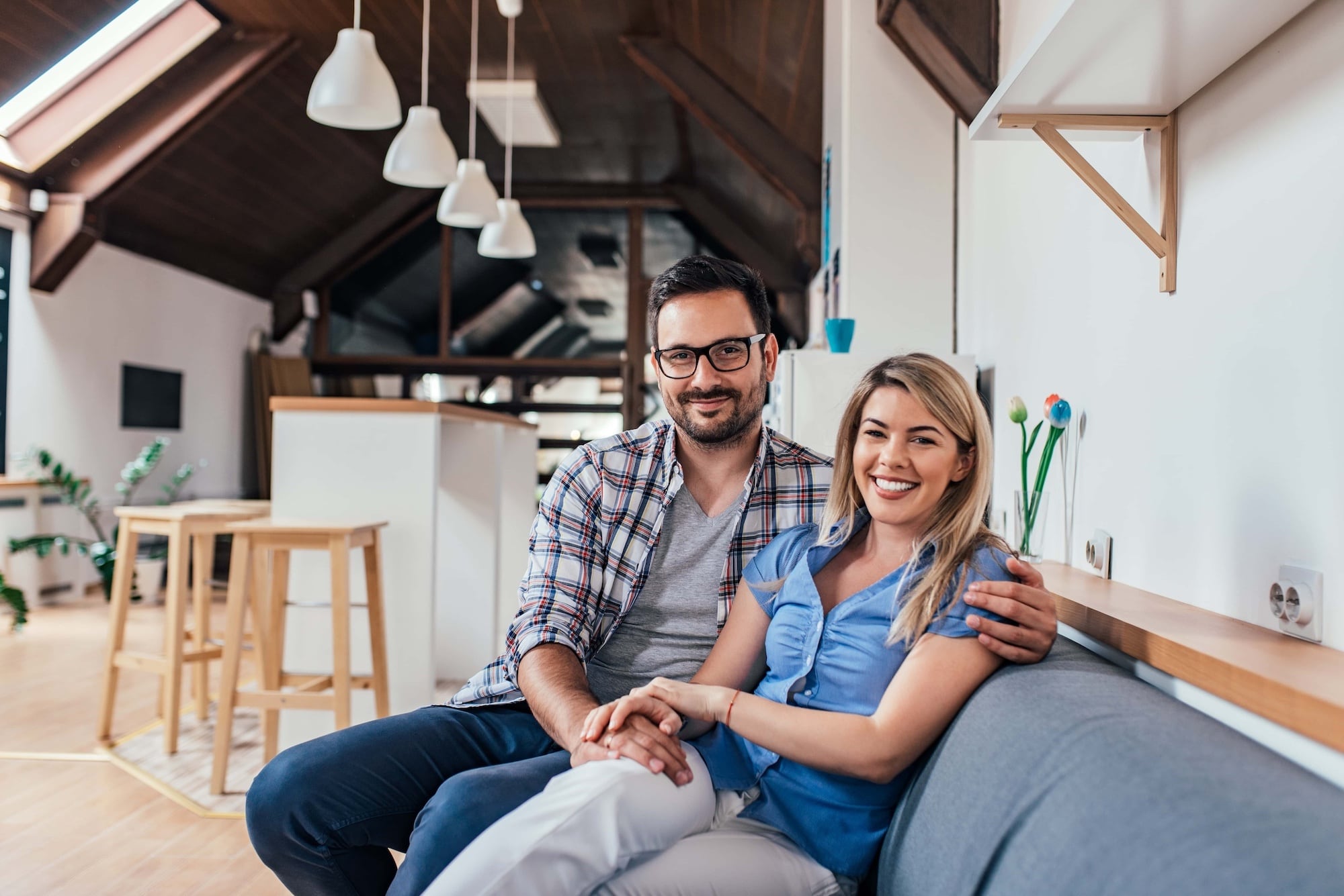 A smiling couple sits on a sofa in a modern, well-lit living room with wooden accents. The man wears glasses and a plaid shirt, while the woman is dressed in a blue top and white pants. Pendant lights and stools are seen in the background.