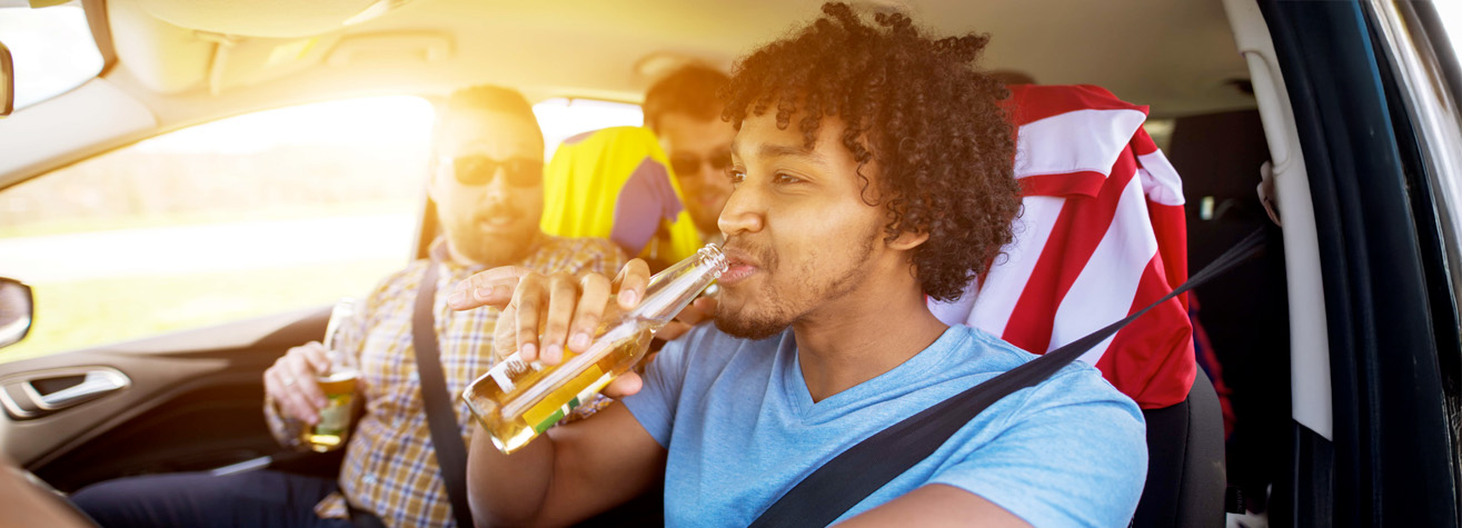 A young man in a blue shirt drinks from a bottle while driving a car. Two passengers in the backseat, with colorful shirts and sunglasses, also hold bottles. The sun is shining brightly through the window.