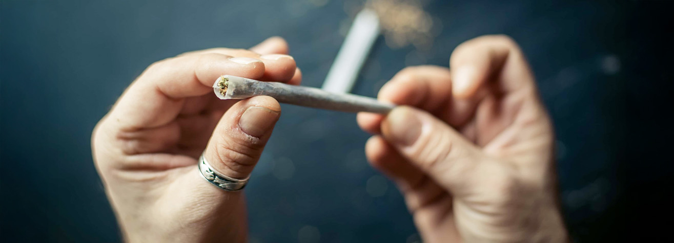 A person holding a rolled cigarette or joint with both hands. The background is out of focus, showing a blurred dark surface. The person is wearing a ring on one finger.