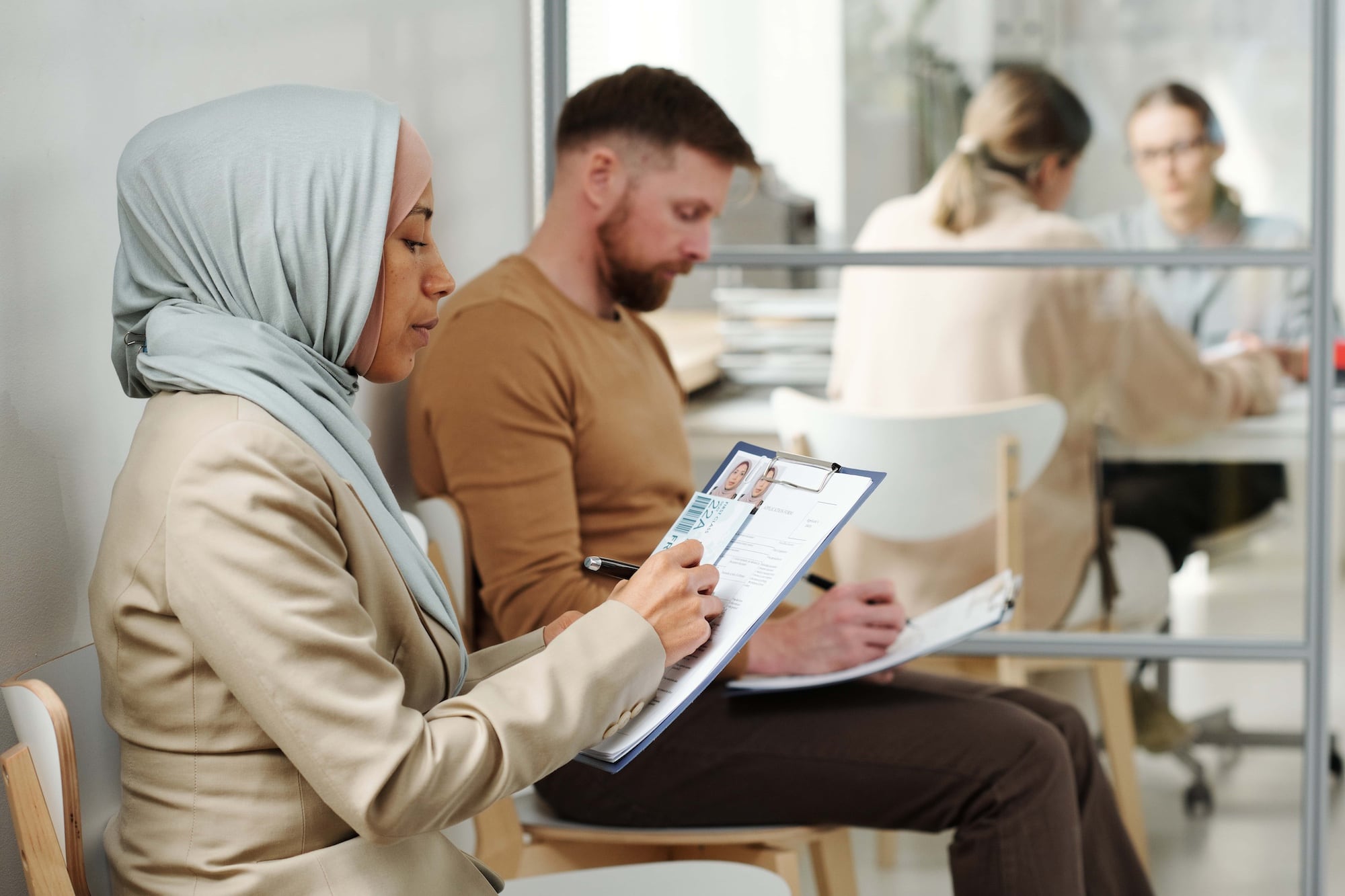 Two people sitting in a waiting room filling out forms. A woman in a hijab writes on a clipboard in the foreground. A man with a beard writes on a form in the background. Two people have a discussion behind a glass wall.