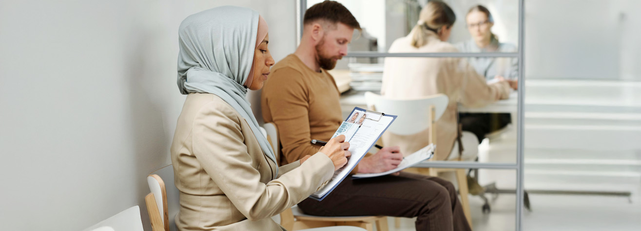 A woman in a light blue hijab and beige blazer writes on a clipboard while sitting in a waiting area. A man in a brown shirt sits beside her, holding a document. Two people are visible in the background through a glass partition.