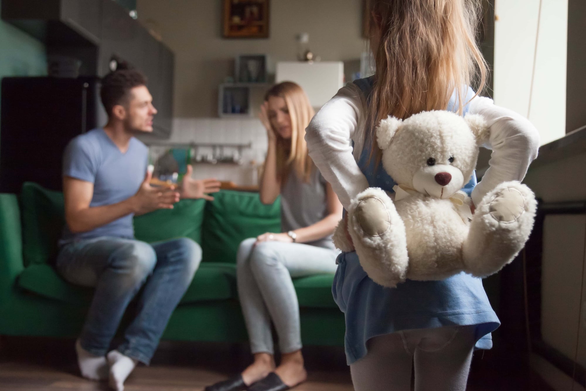 A young girl stands with her back to the camera, holding a teddy bear. In the background, a man and woman sit on a green couch having a serious conversation. The scene appears tense and focused on the seated couple.