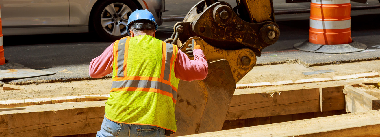Construction worker in a yellow vest and blue helmet stands near a large excavator bucket at a street construction site. Traffic barrels and a car are visible in the background on a sunny day.