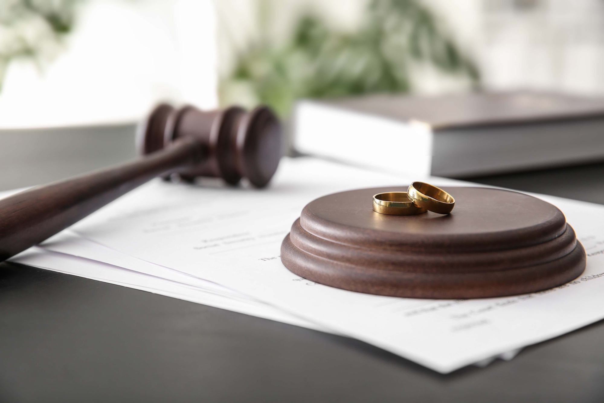 A wooden gavel rests on a round base next to two gold wedding rings on a pile of legal documents. The background is blurred, creating a soft focus on the items, suggesting a divorce or legal proceedings.