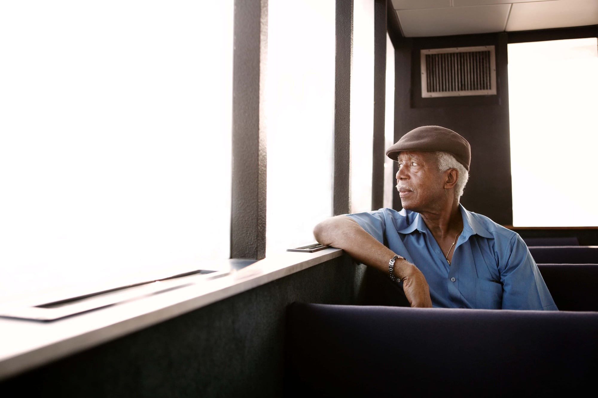 Elderly man in a flat cap and blue shirt sits by a window, gazing outside. Bright light streams in, illuminating his thoughtful expression, as he relaxes in a quiet room with dark interiors.