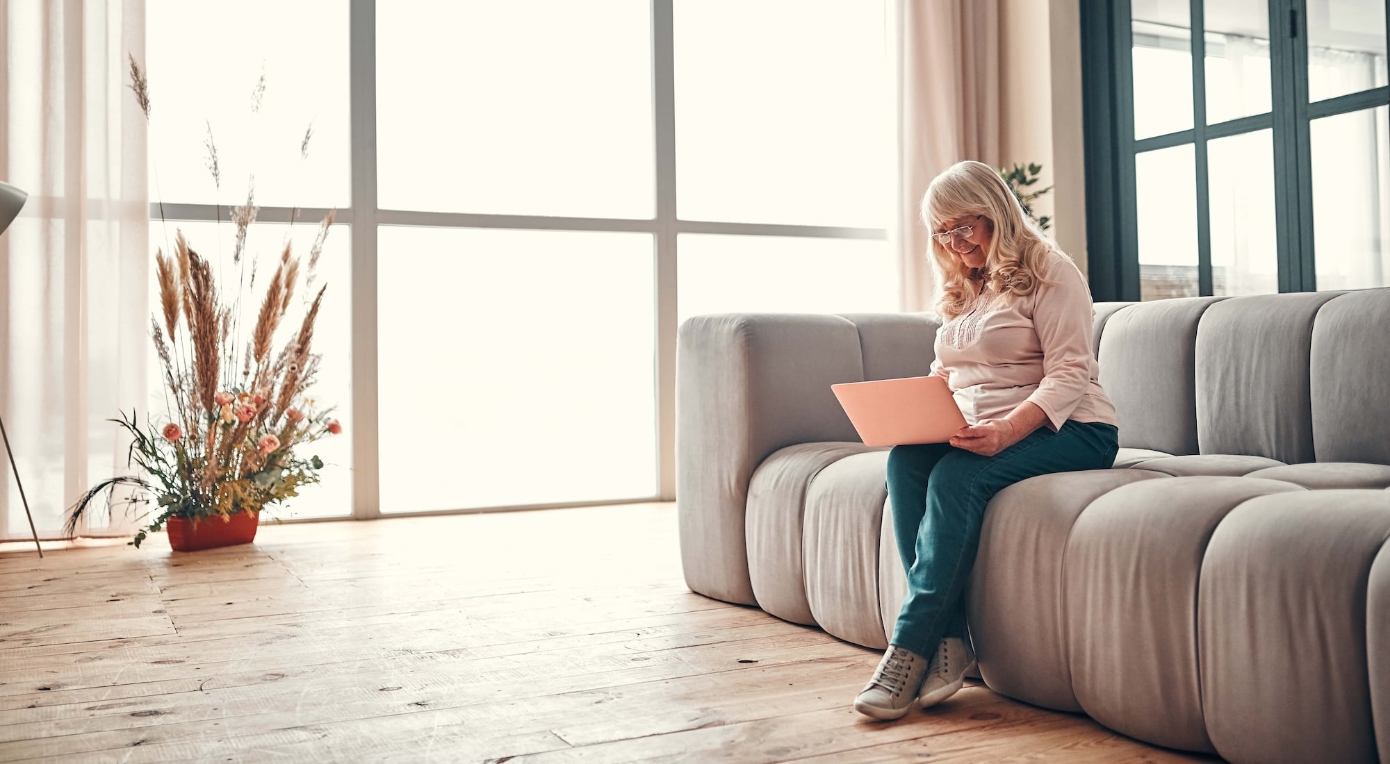 An older woman with long gray hair sits on a gray sofa, using a laptop. She wears glasses, a light shirt, and jeans. The room is bright, with large windows and a potted plant with tall grasses on a wooden floor.