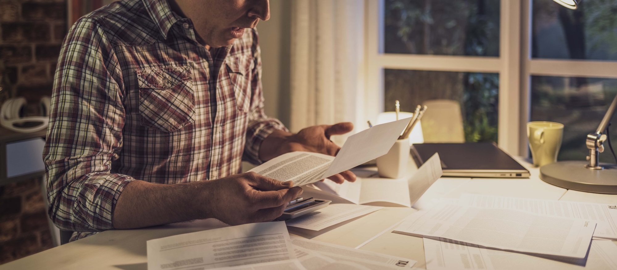 A person in a plaid shirt is sitting at a desk, intently reading a document among scattered papers. A laptop, pen, and coffee cup are on the desk, and a window with a tree view is in the background. The room is softly lit.