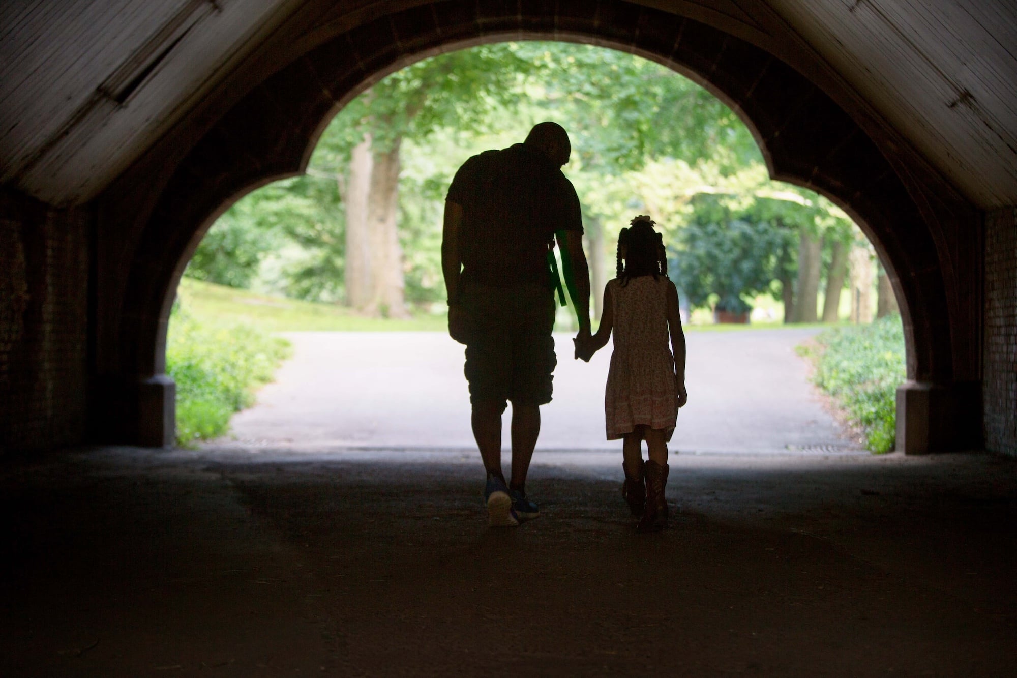 A silhouette of a man and a girl holding hands as they walk through a tunnel towards a sunlit park, with greenery visible in the background. The scene conveys a sense of bonding and tranquility.