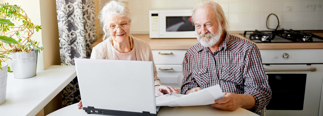 Elderly couple sitting at a kitchen table using a laptop. The woman wears a light sweater, and the man has a plaid shirt. They appear to be looking at documents. The kitchen has white cabinets and a microwave. There's a potted plant nearby.