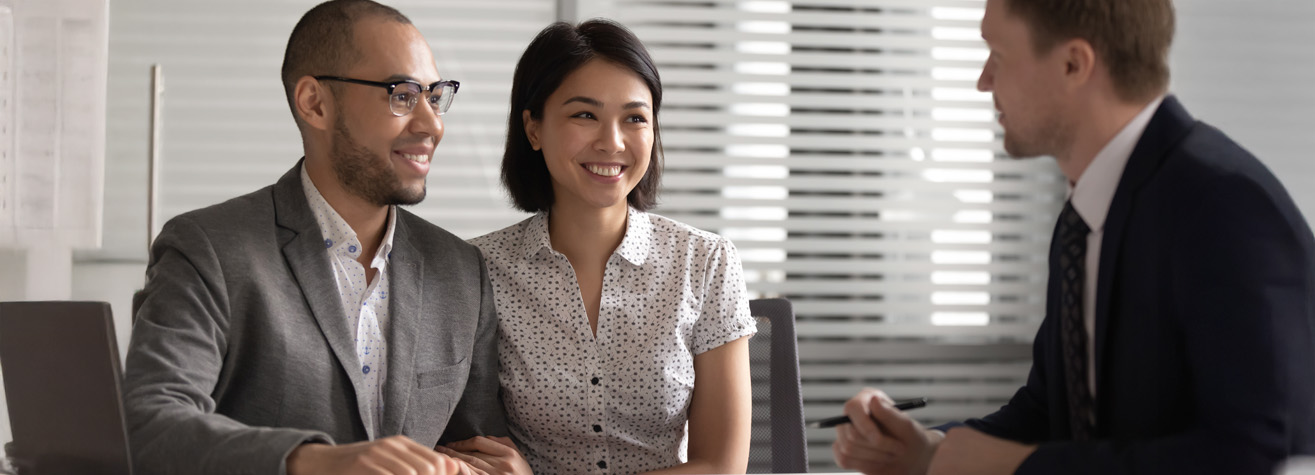 A couple sits across from a professional who is holding a pen. The couple appears engaged and happy, while the professional, in a suit, seems to be explaining something. They are in an office setting with blinds in the background.