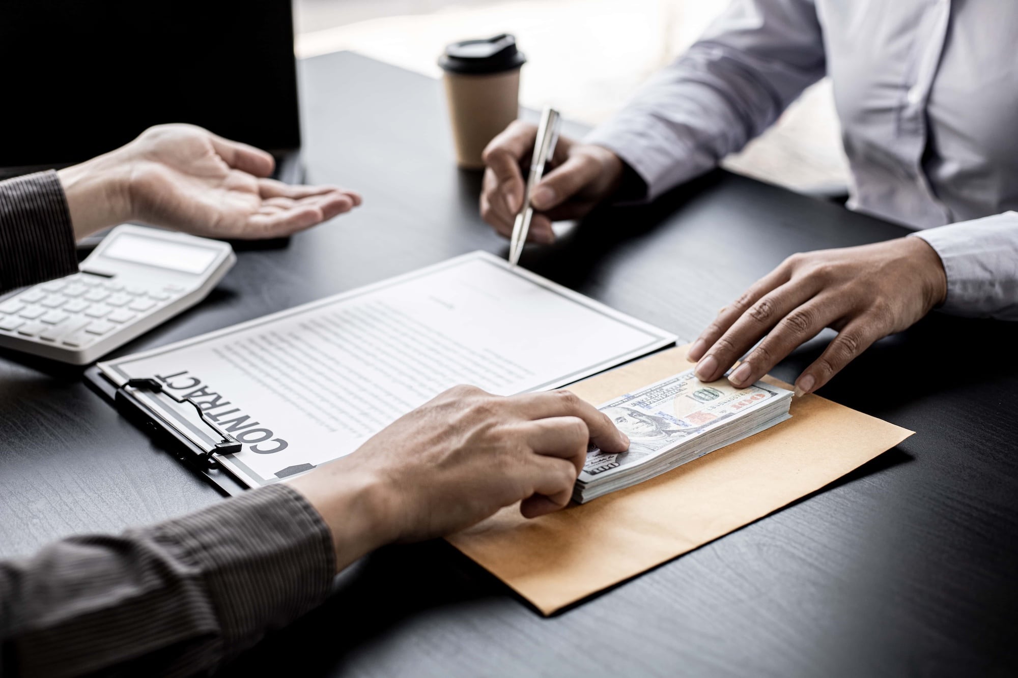 Two people are sitting at a table. One hands money in an envelope to the other, who is signing a contract with a pen. A calculator and a coffee cup are on the table.