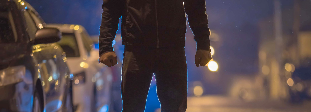 Silhouette of a person standing in the middle of a dimly lit street at night, with hands clenched into fists. Cars are parked on either side, and streetlights illuminate the background with a soft, blurry glow.