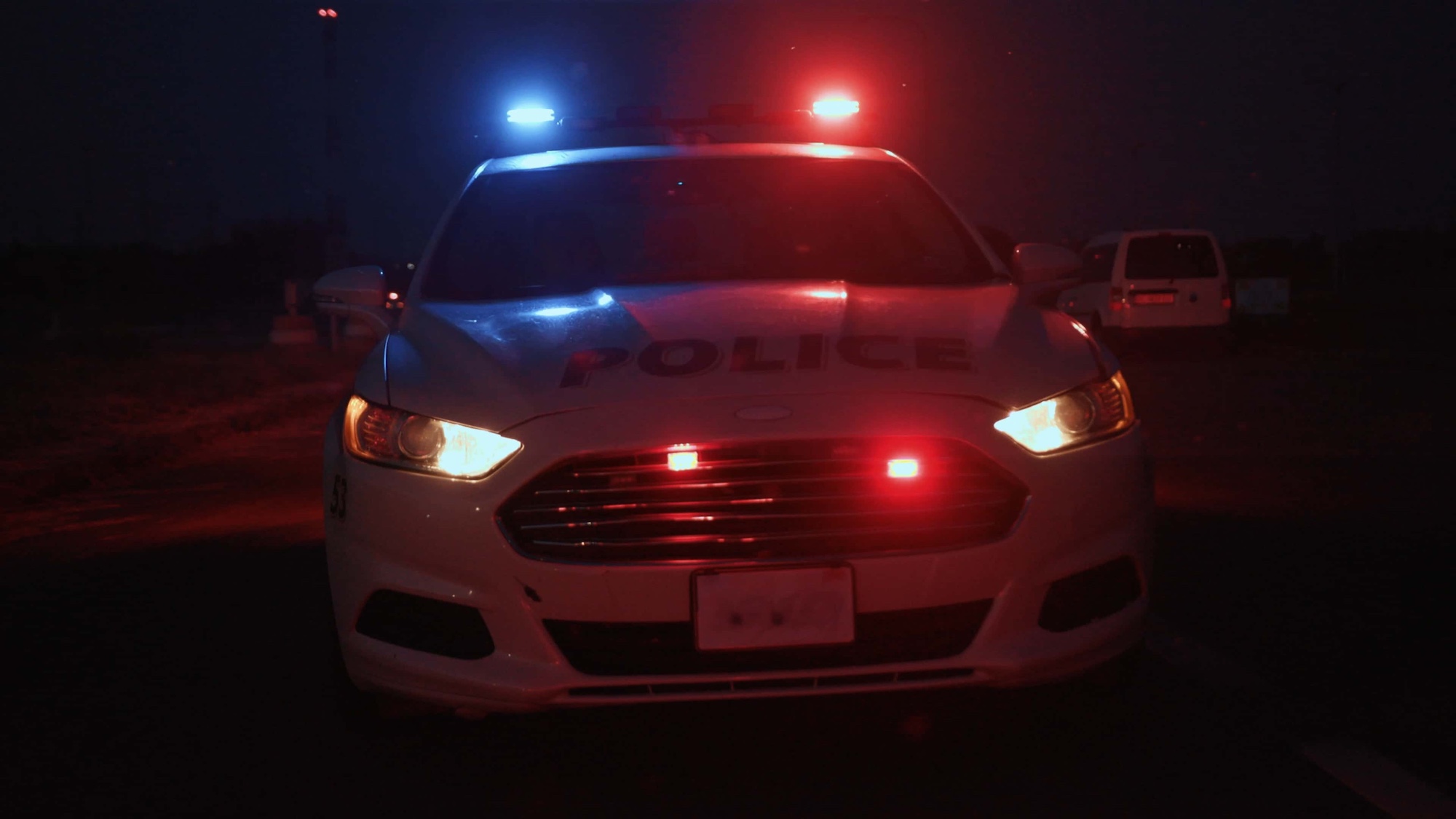 A police car at night with flashing red and blue lights, parked on a dark road. The front headlights are on, illuminating the area ahead. Another vehicle is visible in the background.