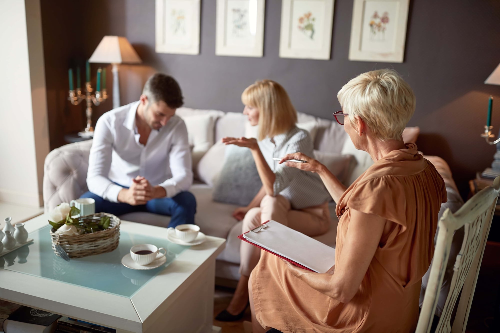 A couple sits on a sofa, engaging in conversation with an older woman holding a clipboard. The room is cozy, decorated with framed art and a lit lamp. A coffee table is in the foreground, featuring cups, a teapot, and a basket.