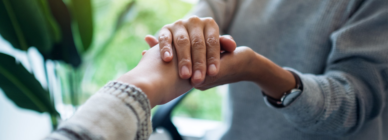 Two people are holding hands gently. One person is wearing a gray sweater, and the other wears a striped top. The background is softly blurred with green tones, suggesting plants. The scene conveys a sense of support and compassion.