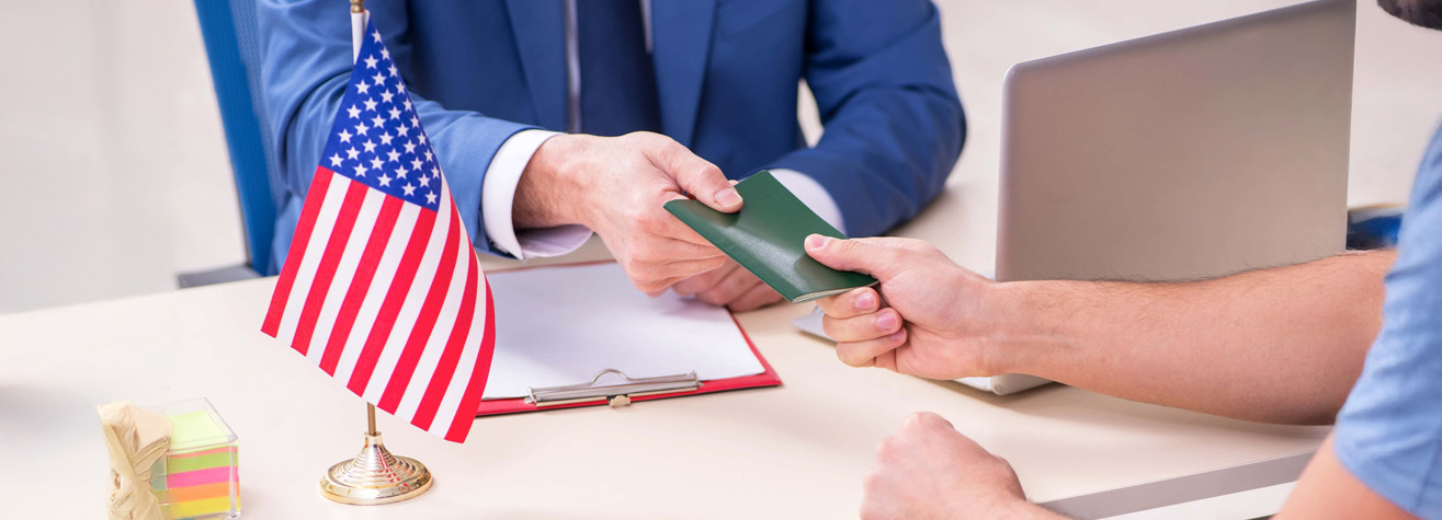 Two people exchange a green passport across a desk with a laptop and small U.S. flag. One person wears a blue suit, while the other has a bare arm. A clipboard with papers is also visible on the desk.