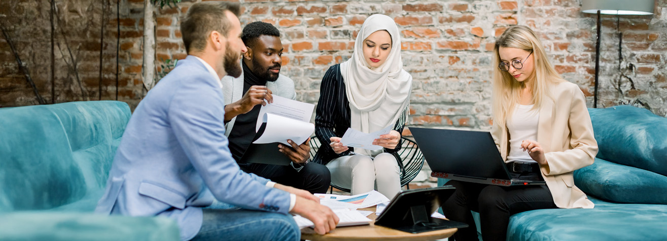 Four people are seated on sofas in a casual office setting, engaged in a discussion. They are reviewing documents and working on laptops. The room has brick walls and a relaxed atmosphere.