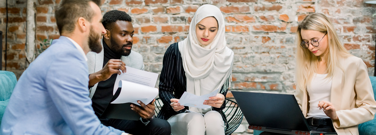 Four people sit together in a modern office setting with brick walls. They are engaged in discussion, holding papers and using a laptop. The group is diverse, featuring different backgrounds and styles.