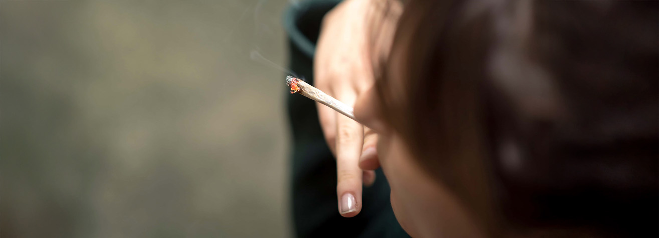 A person is holding a lit cigarette or joint close to their mouth. The focus is on the hand and the smoking item, with some smoke visible. The background is blurred.