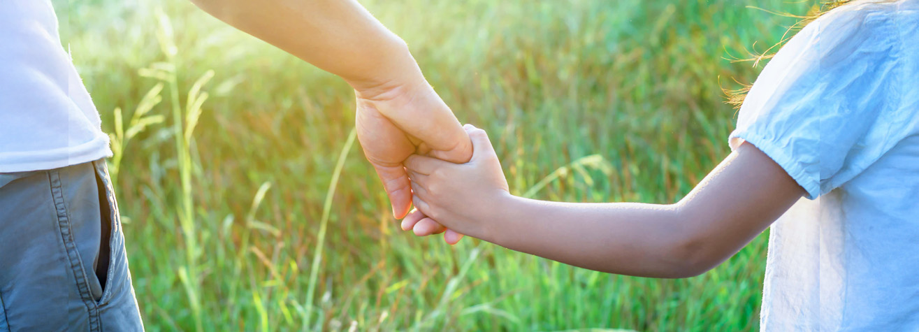 An adult and a child are holding hands, standing in a sunlit field of green grass. The child is in a white shirt, and the adult is in a white shirt and brown pants. The scene is warm and serene, with sunlight filtering through the grass.