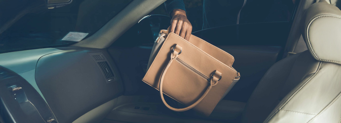A person reaching into a car to place or retrieve a tan leather handbag from the passenger seat. The interior of the car is beige and the scene is slightly dimly lit.