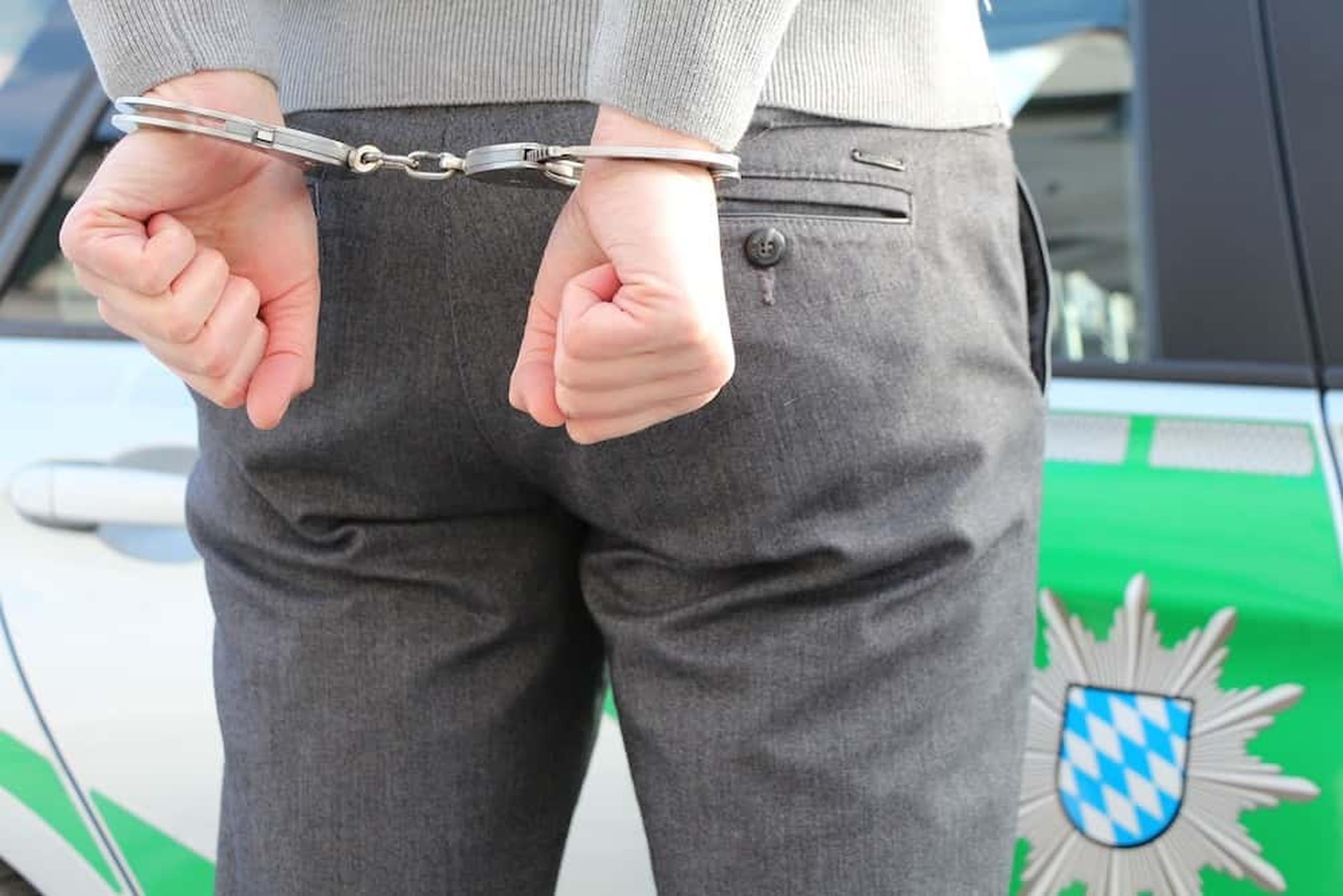 A person in gray suit pants is handcuffed with their hands behind their back. They are standing in front of a police car, which has a green and white design with an emblem featuring a blue and white checkered shield.