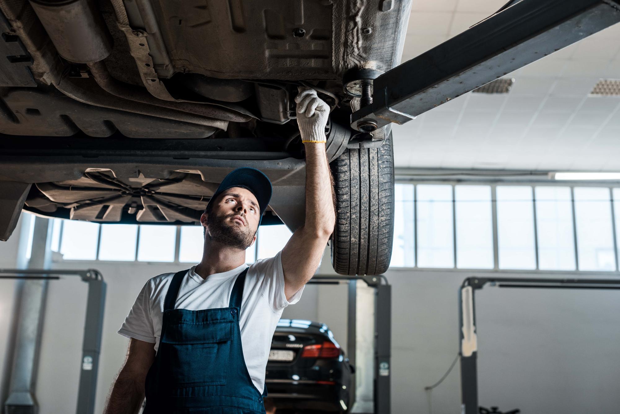Mechanic wearing a cap and overalls inspects the underside of a lifted car in a garage. He is looking up while working, with tools in the background. The garage is well-lit with a window in the upper part of the image.