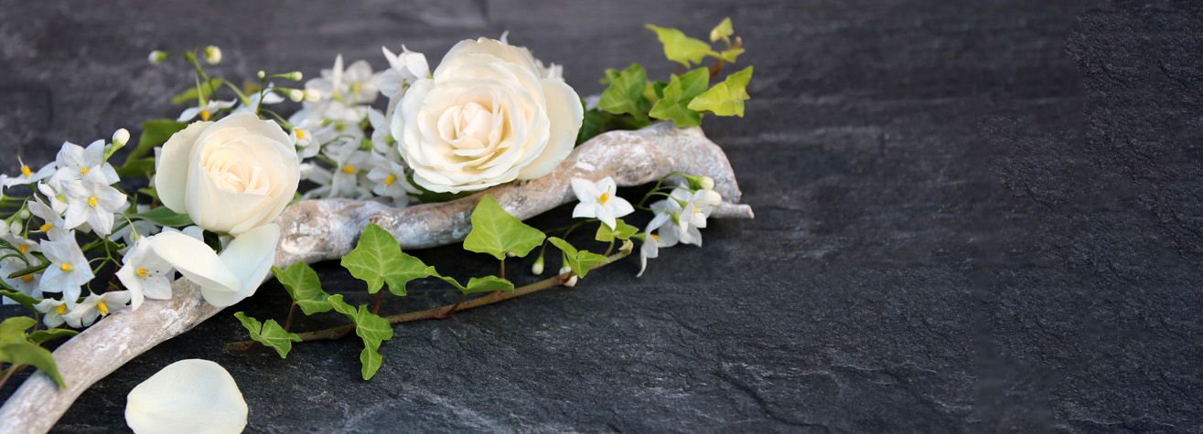 White roses and small white flowers with green leaves rest on a twisted branch against a dark, textured stone background. A few petals are scattered nearby.