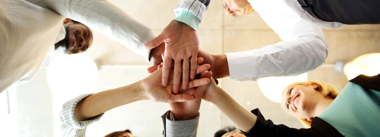 A diverse group of people standing in a circle, each reaching into the center to place one hand on top of the others. They are indoors, and the photo is taken from below, looking upwards. The mood feels positive and collaborative.