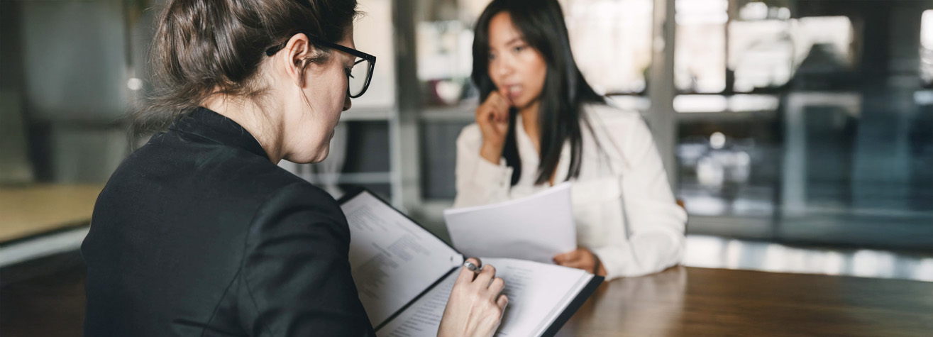 Two women in professional attire are sitting across a table from each other in a modern office setting. One is holding a folder and pen, appearing to discuss documents with the other, who listens attentively while holding a paper.